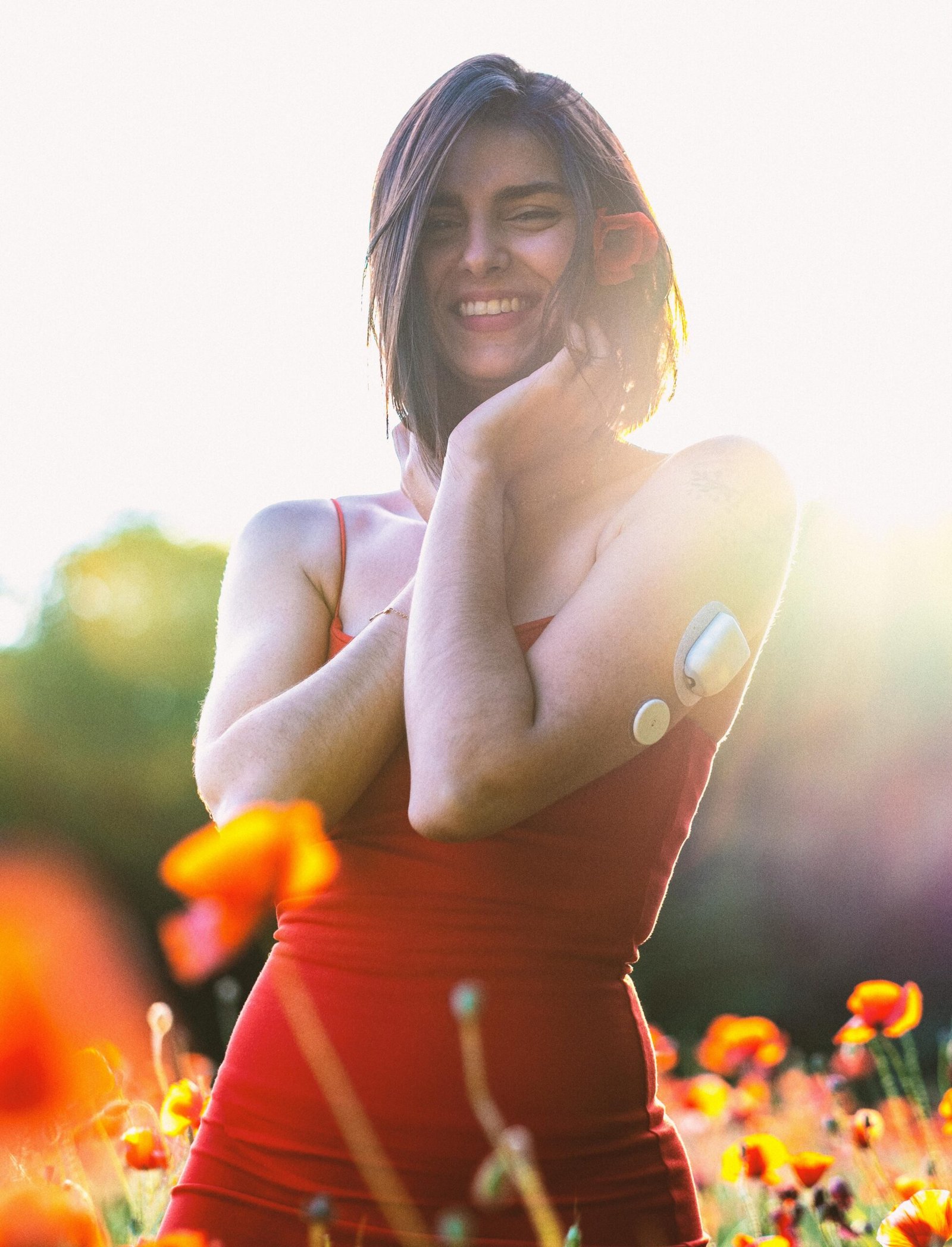 A joyful woman with a diabetes monitor stands in a vibrant flower field, embracing the sunset.